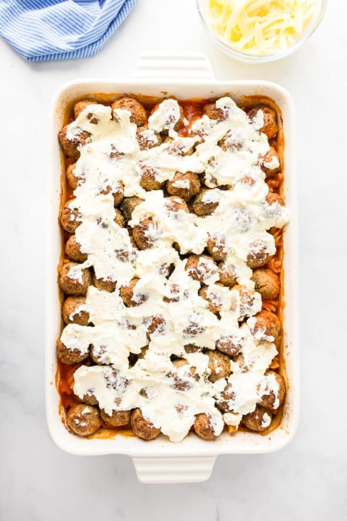 A rectangular baking dish filled with cooked meatballs covered in melted cheese sauce, set on a white surface. A blue cloth and a bowl of shredded cheese are nearby.