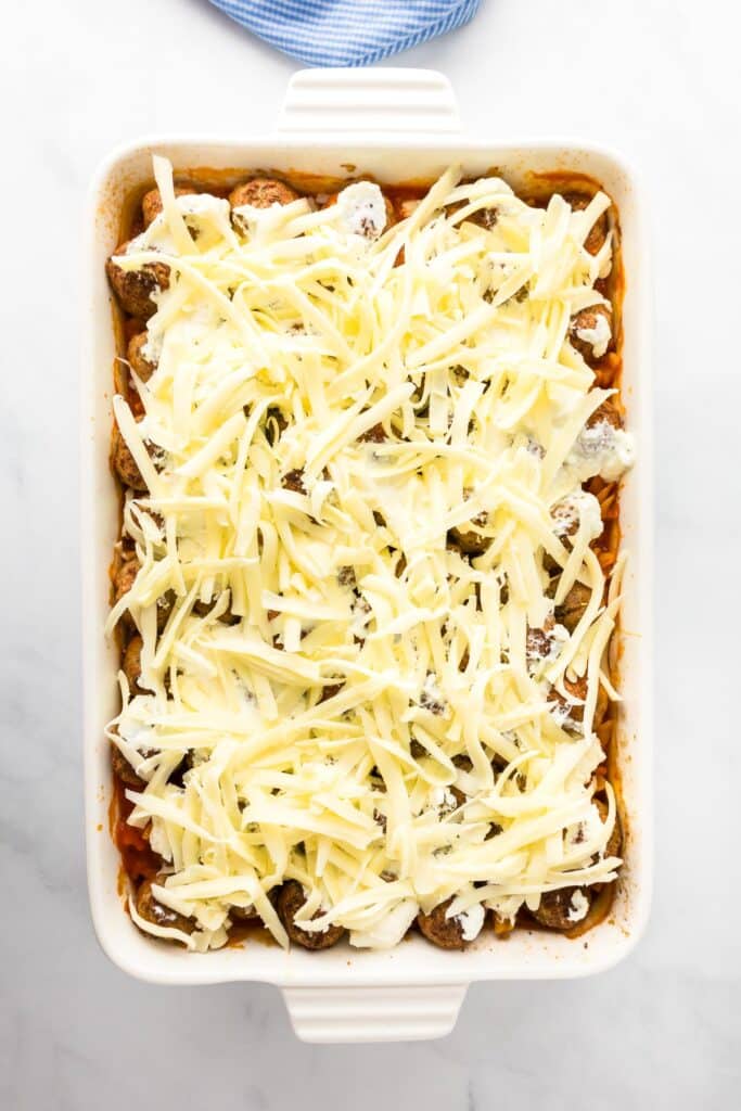 A rectangular casserole dish filled with unbaked meatballs, tomato sauce, and topped with grated cheese on a marble surface.