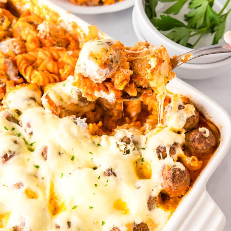 Square view of dump and bake meatball casserole with pasta, meatballs and melted cheese in a baking dish being served with a spoon.