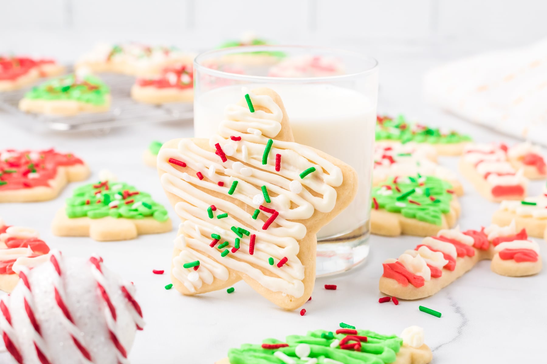 A star-shaped cut out sugar cookie cookie with white icing and colorful sprinkles leans against a glass of milk. Other decorated cookies are on the table.