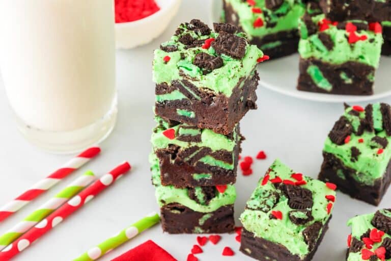 Stack of mint chocolate Grinch brownies with green frosting, topped with chocolate pieces and red heart sprinkles, next to colorful straws and a glass of milk.