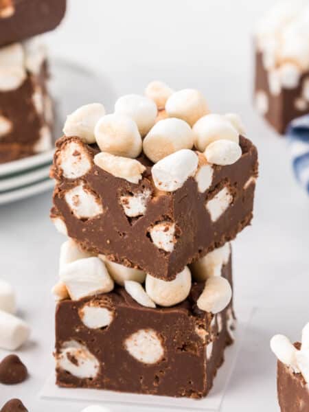 Stacked hot chocolate chocolate fudge pieces with marshmallows on a white counter, surrounded by more pieces of marshmallow topped chocolate fudge.