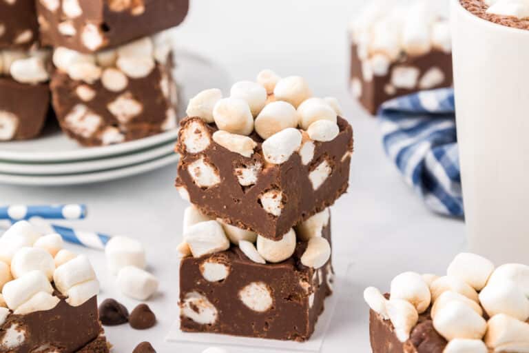 Stacked hot chocolate chocolate fudge pieces with marshmallows on a white counter, surrounded by more pieces of marshmallow topped chocolate fudge.