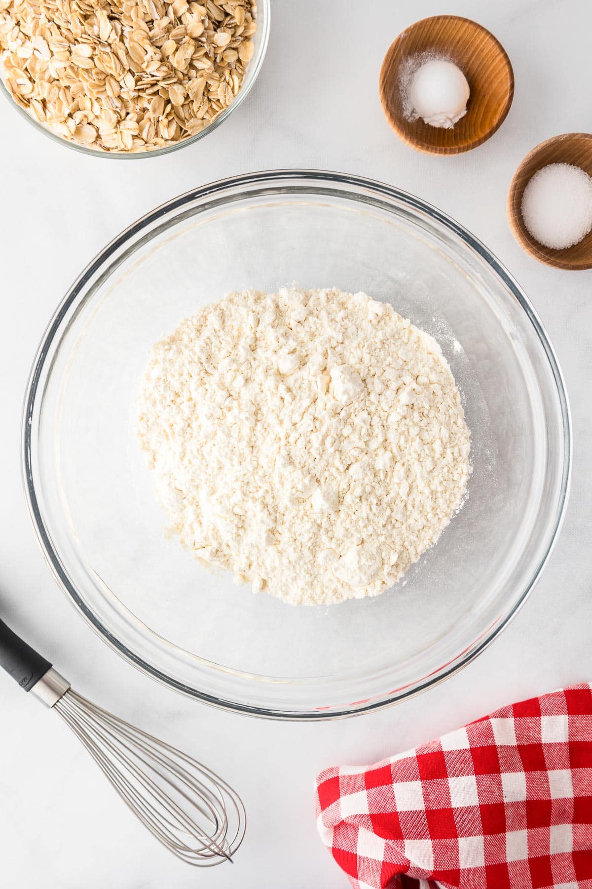 A glass bowl with flour, surrounded by oats, salt, and sugar in bowls to mix for oatmeal raisin cookies.