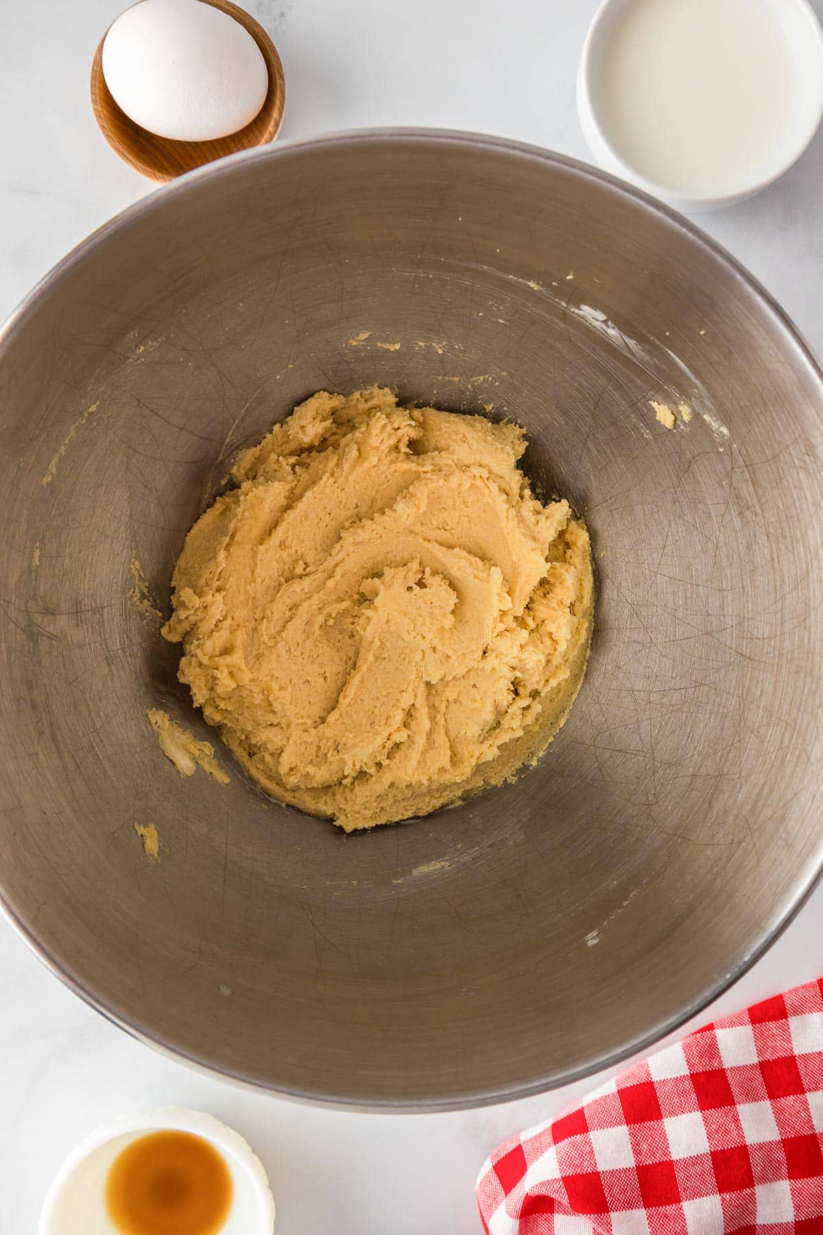 A metal bowl with butter and sugar mixed together with an egg, vanilla and milk in bowls nearby for oatmeal raisin cookies.