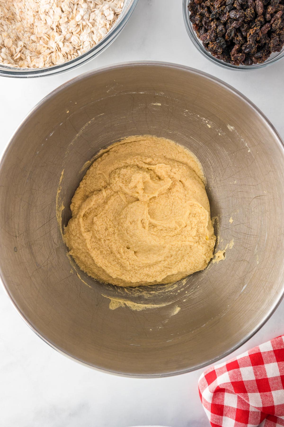 A metal bowl contains cookie dough before adding oats and raisins which are nearby in bowls