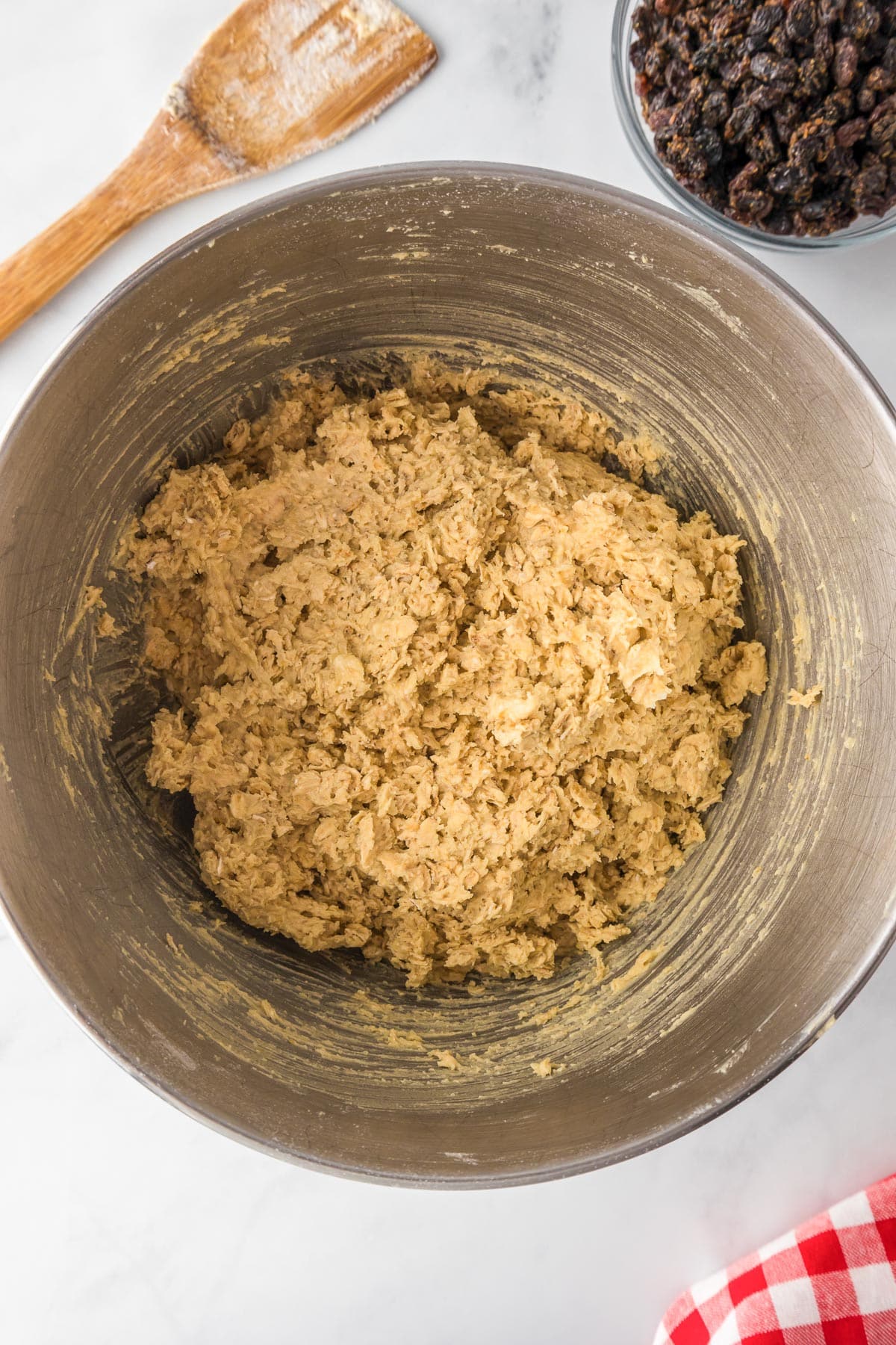 A mixing bowl filled with oatmeal cookie dough on a counter with a bowl of raisins nearby for oatmeal raisin cookies.