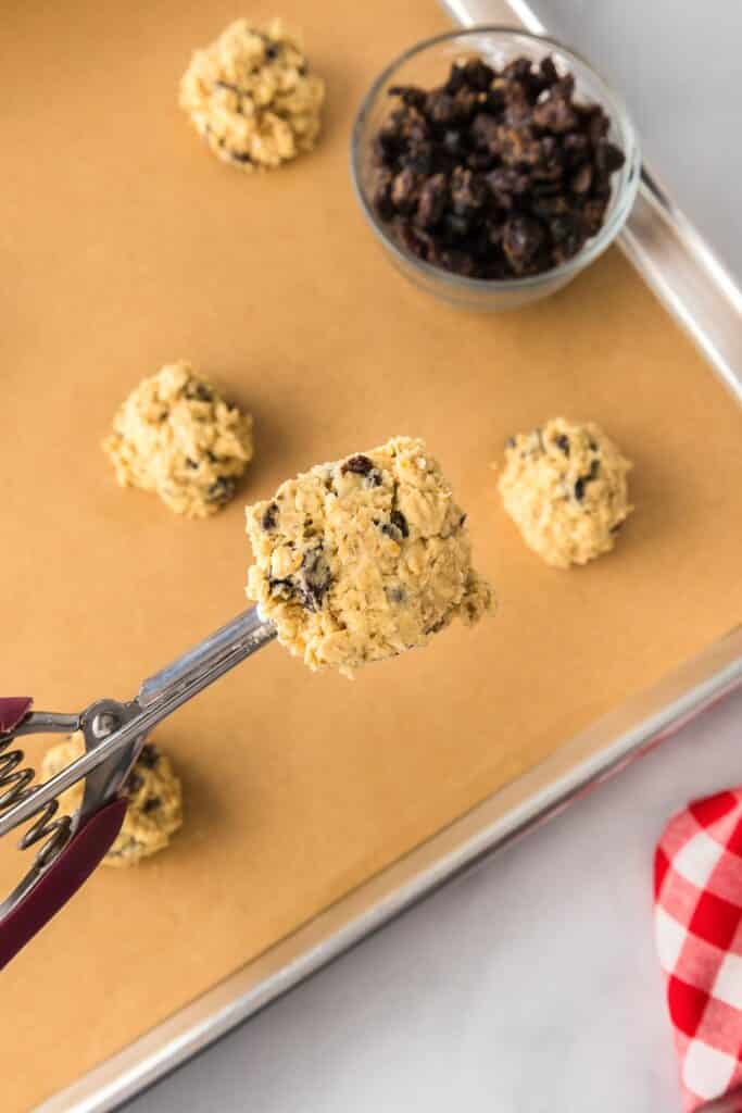 Oatmeal raisins cookie dough being scooped with a cookie scoop into cookie dough balls on a pan lined with parchment paper with a bowl of raisins nearby.