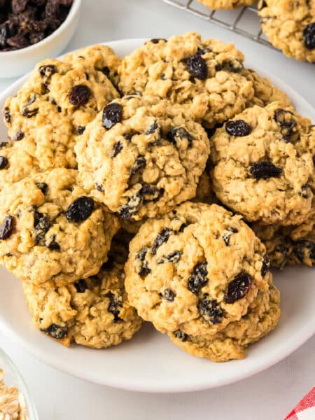 A plate of homemade oatmeal raisin cookies with a bowl of raisins and oats nearby.