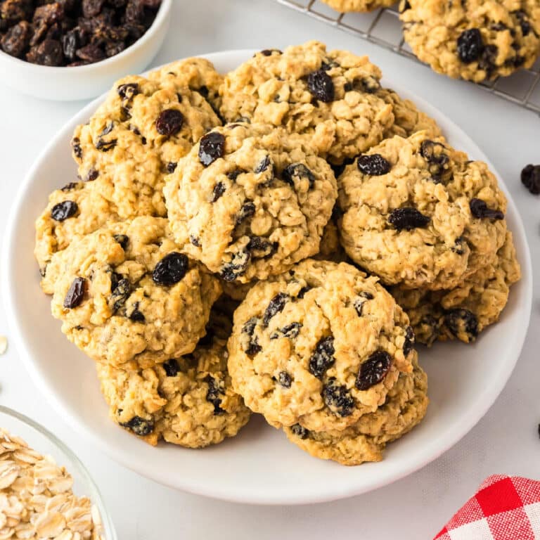 A plate of homemade oatmeal raisin cookies with a bowl of raisins and oats nearby.