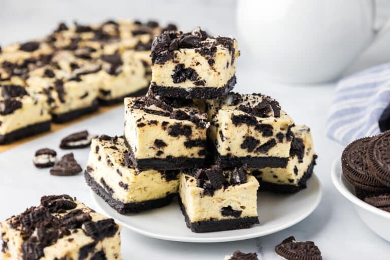 A plate of cookies and cream cheesecake bars stacked neatly, topped with crumbled Oreo chocolate cookies with more cheesecake bars in the background on the counter.