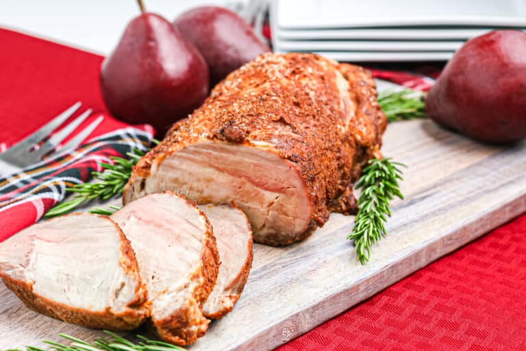 Sliced roast pork loin roast on a wooden board with red pears and rosemary garnish.