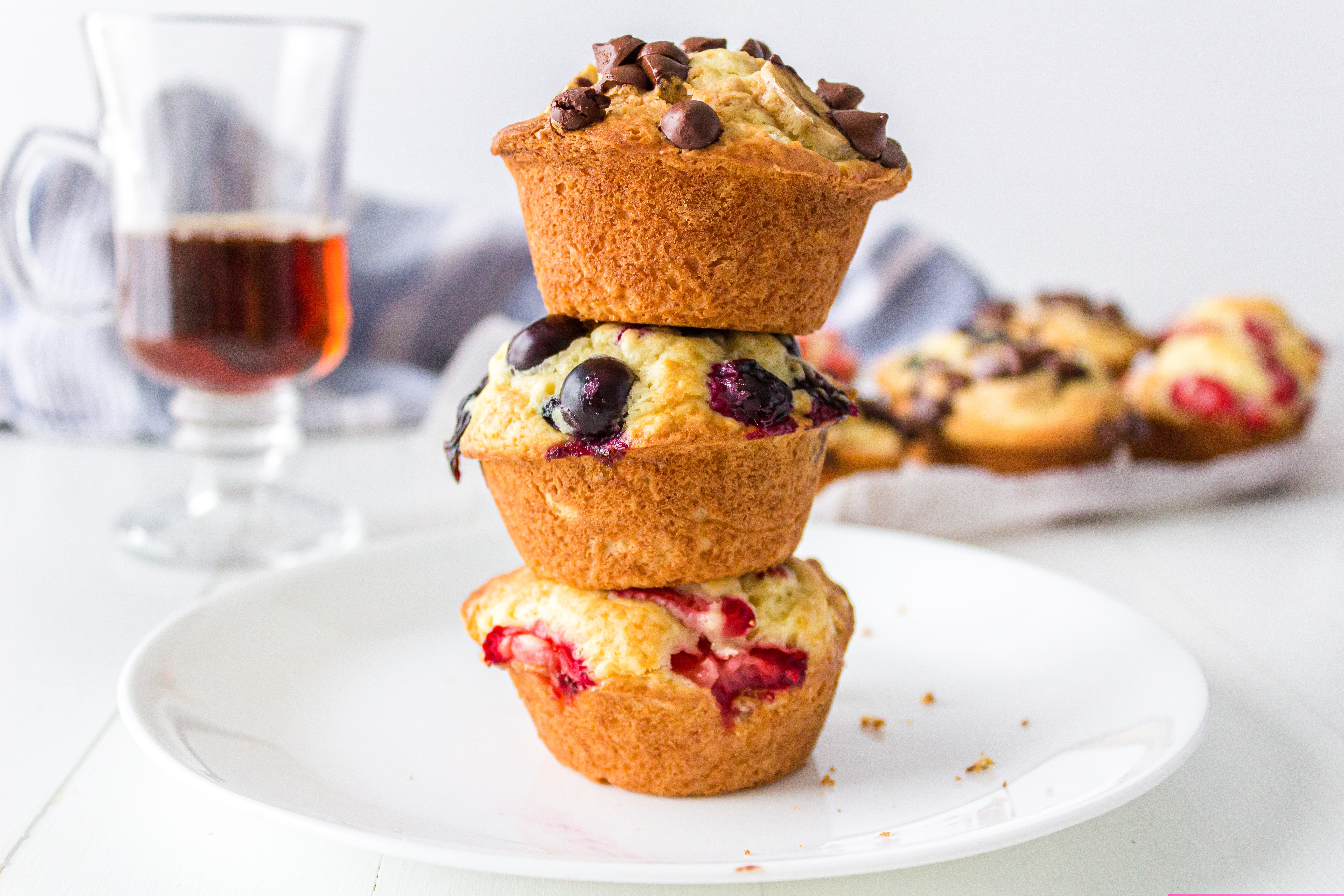 Three muffins stacked on top of each other on a plate, one with chocolate chips, one with blueberries and one with strawberry pieces.