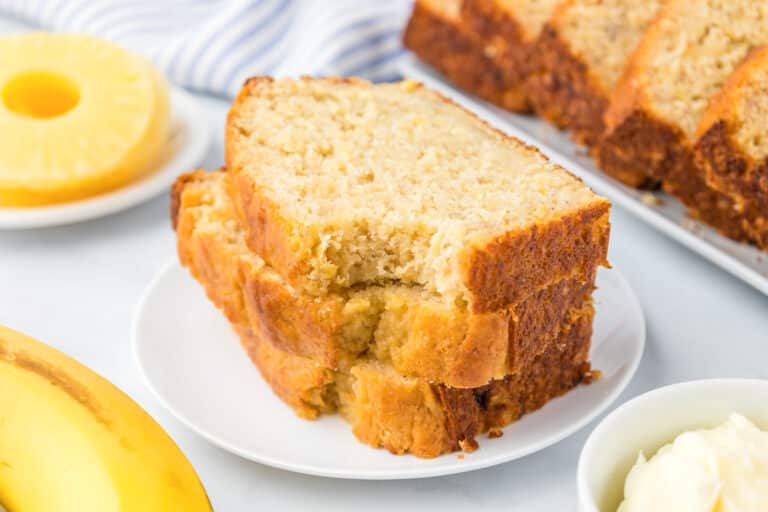 Slices of pineapple banana bread stacked on a plate missing a bite from the top piece, with pineapple and banana nearby on the counter.