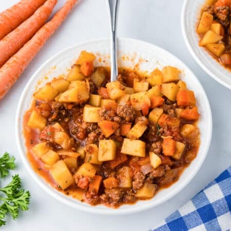 A white bowl full of hamburger stew with potatoes and carrots on a counter with a spoon with more stew in a bowl and some fresh vegetables nearby.