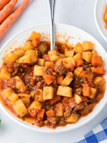 A white bowl full of hamburger stew with potatoes and carrots on a counter with a spoon with more stew in a bowl and some fresh vegetables nearby.