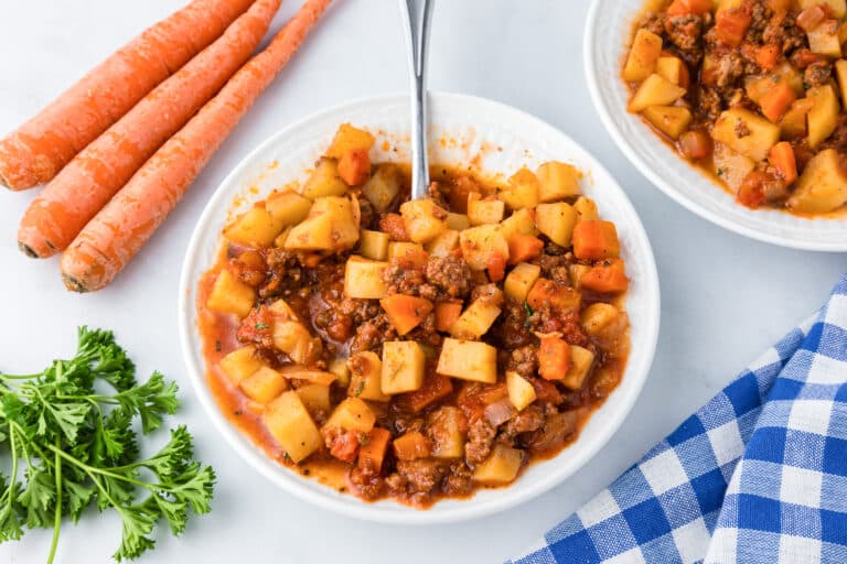 A white bowl full of hamburger stew with potatoes and carrots on a counter with a spoon with more stew in a bowl and some fresh vegetables nearby.