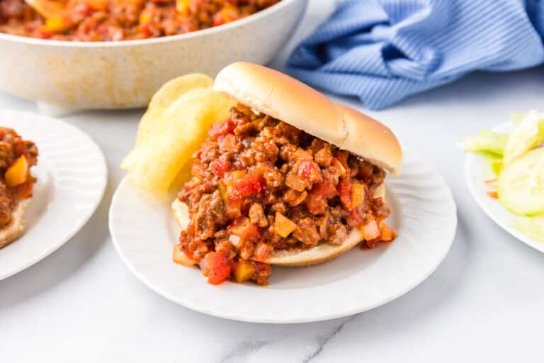 A sloppy joe sandwich on a plate next to potato chips on a counter.