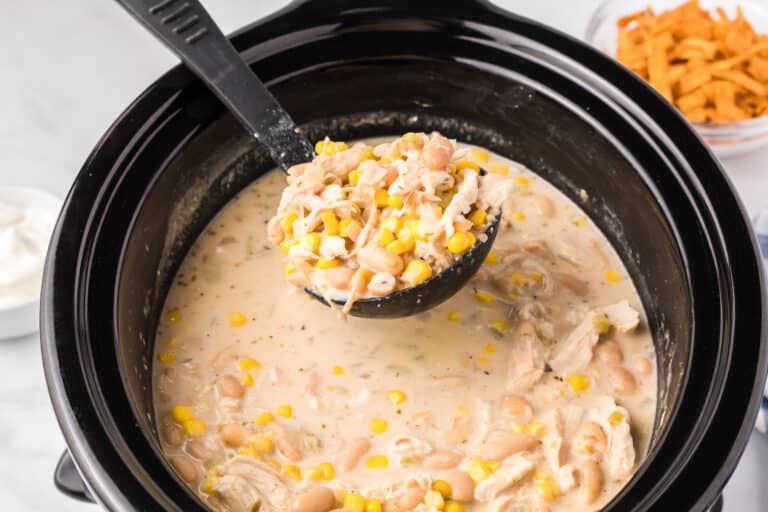 A ladle scooping creamy white chicken chili from a slow cooker full of chicken, white beans, corn, and a creamy broth.