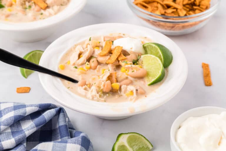 A bowl of creamy white chicken chili topped with tortilla strips and lime slices, with a spoon on a counter with more toppings nearby.