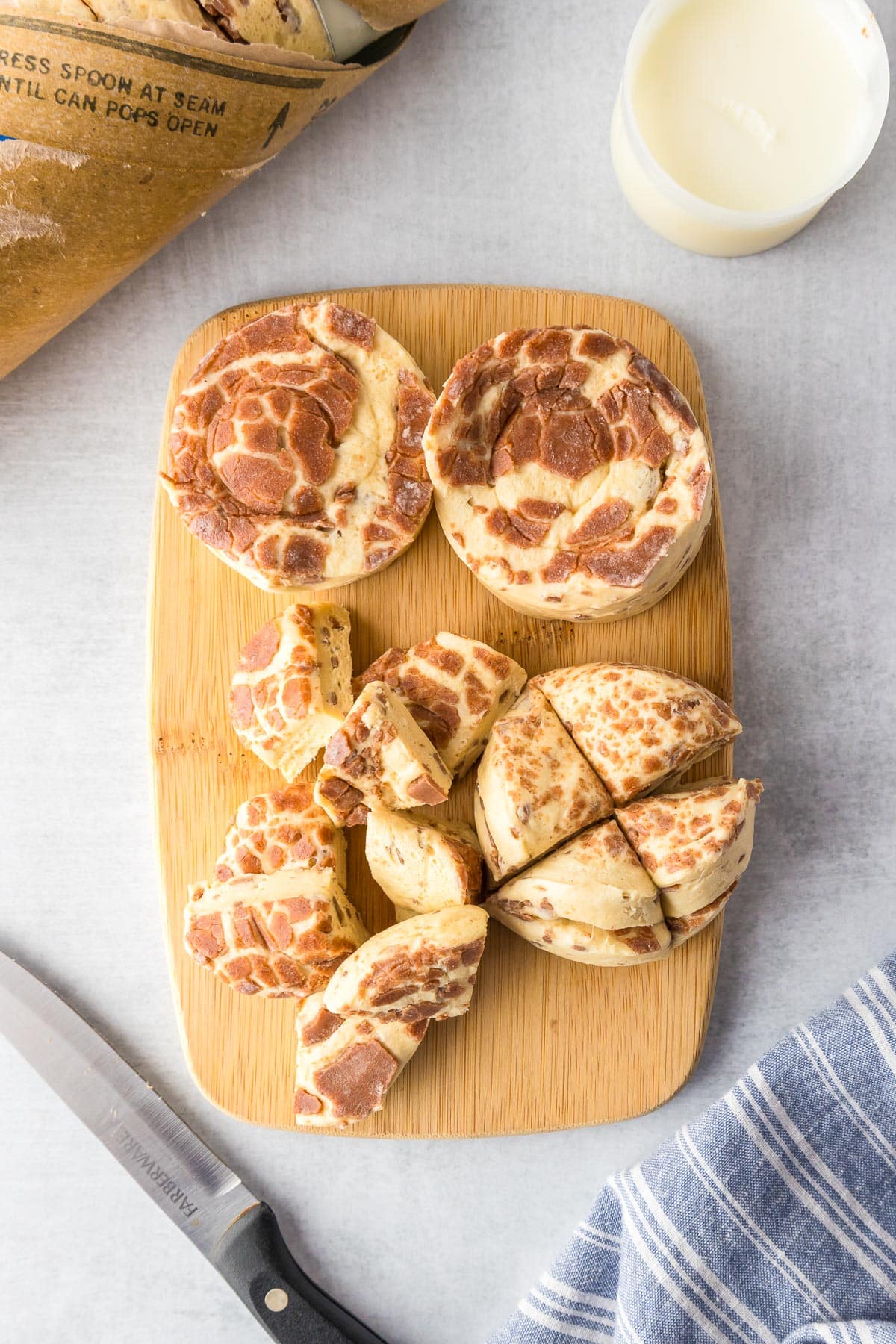 Cinnamon roll dough biscuits from a can being sliced on a cutting board into four pieces for cinnamon roll casserole with the tube, the frosting container and a knife nearby on the counter.