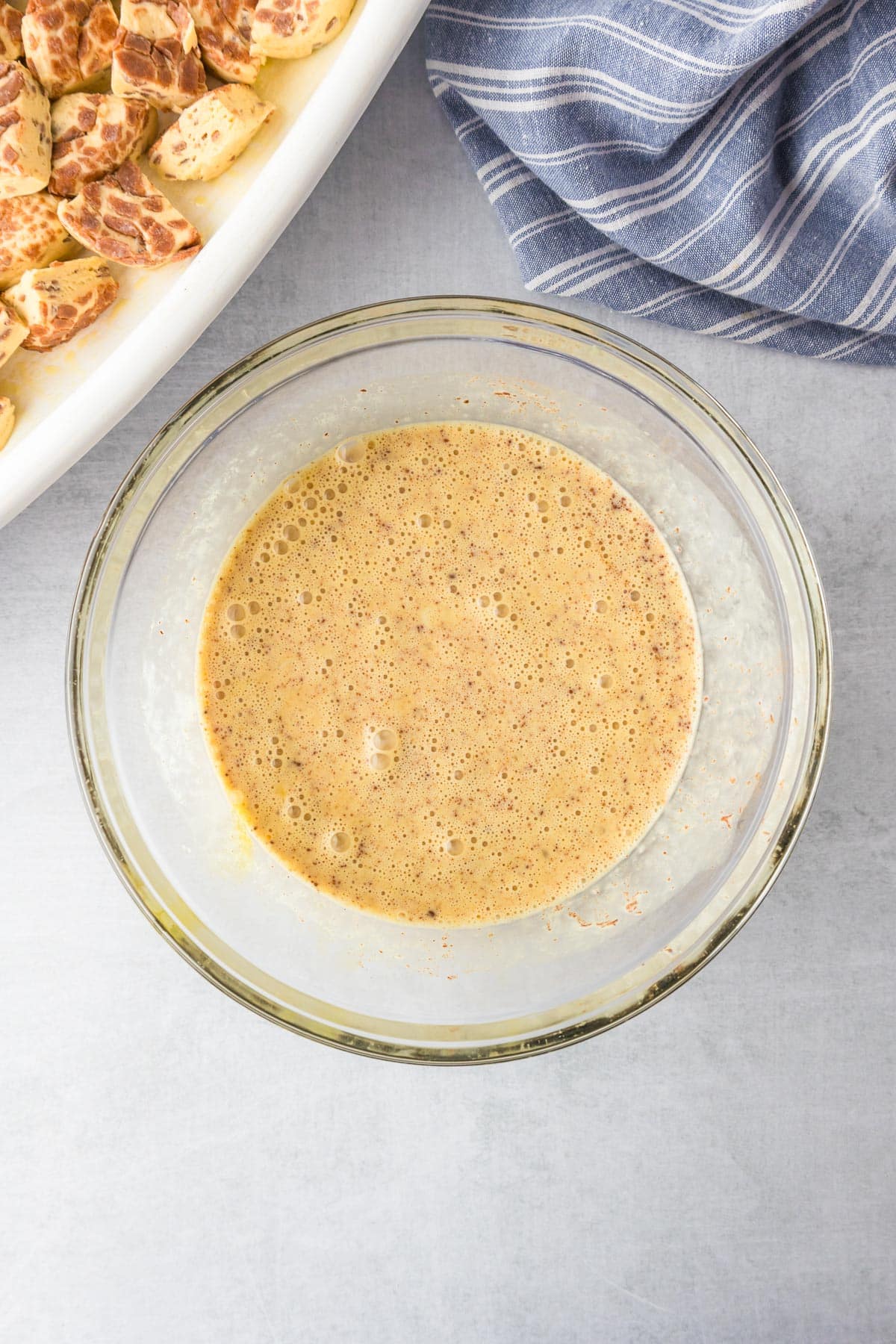 A glass bowl filled with the cinnamon egg custard mixture for cinnamon roll casserole next to a pan full of cinnamon roll biscuit pieces.