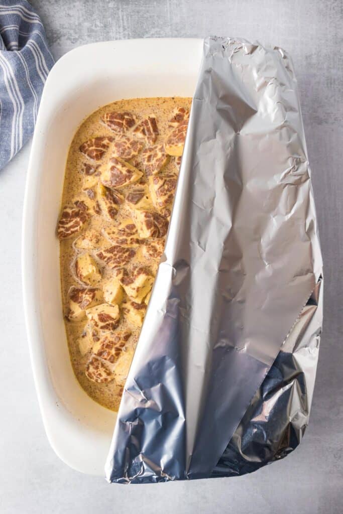 A baking dish with cinnamon roll dough pieces in a custard for cinnamon roll casserole being covered by aluminum foil.