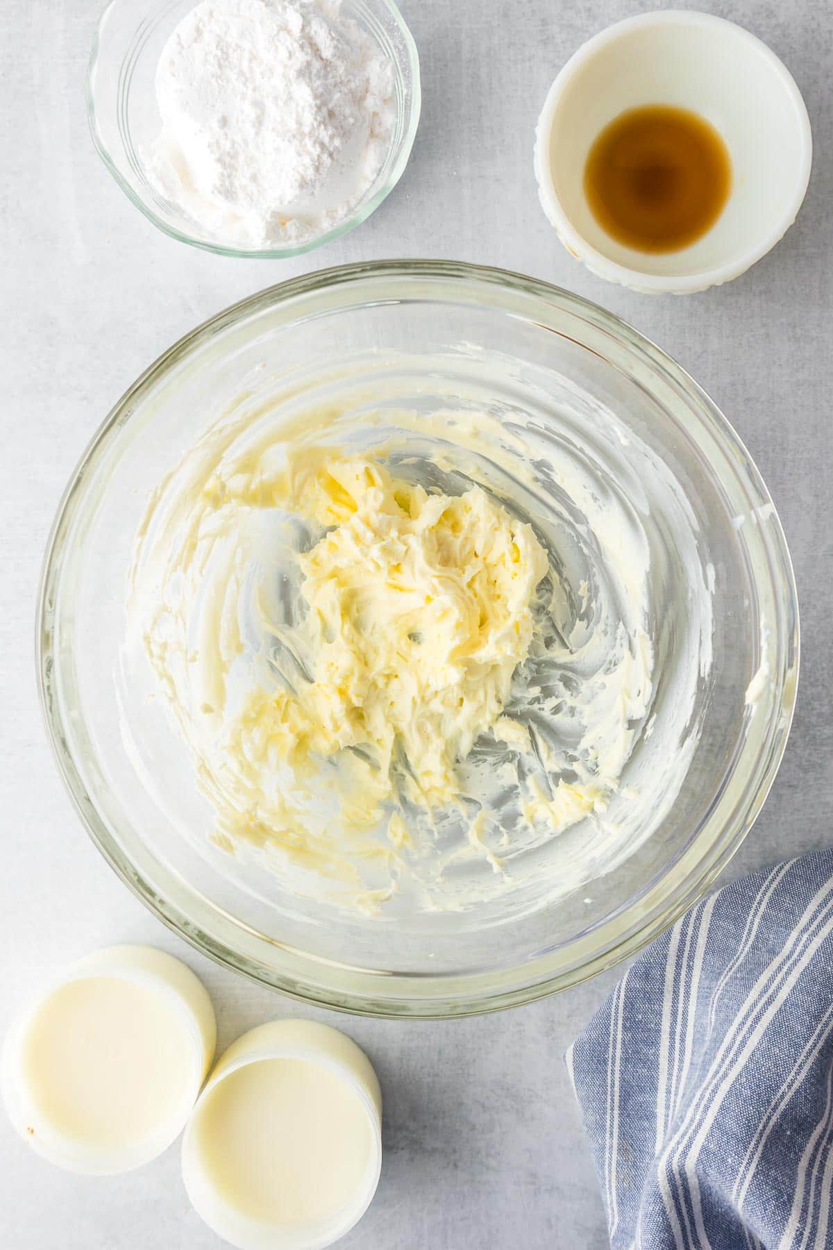 A glass bowl with creamed butter and cream cheese being mixed into frosting surrounded by bowls of powdered sugar, vanilla and two packets of cinnamon roll frosting from the tubes of dough for cinnamon roll casserole.