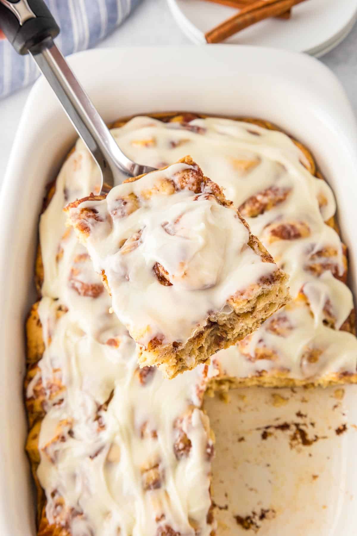 A spatula lifts a frosted cinnamon roll casserole piece from a baking dish filled with more casserole.