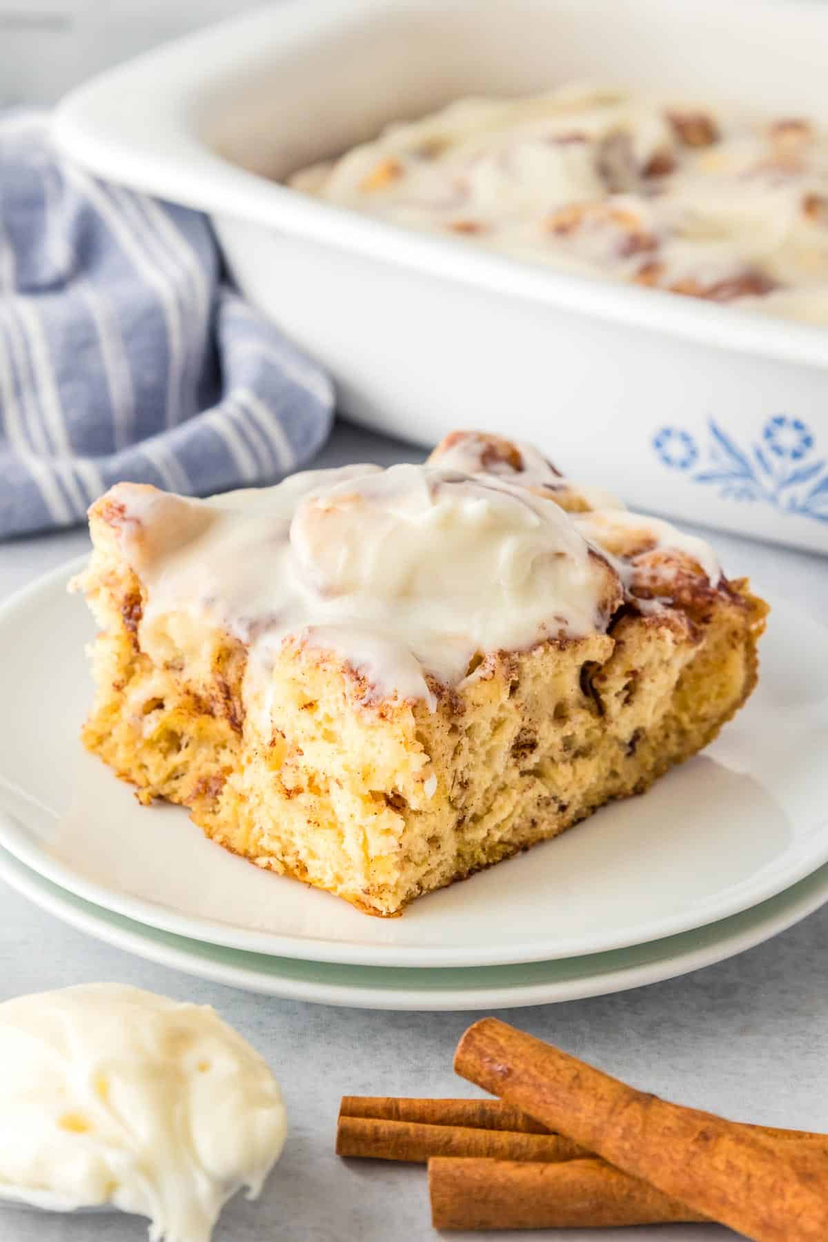 A single frosted cinnamon roll casserole slice square on plate, next to the more cinnamon roll casserole in the pan.