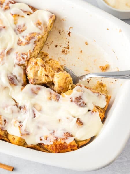 A white baking dish with frosted cinnamon roll casserole, partially served as a spoon scoops from the pan.