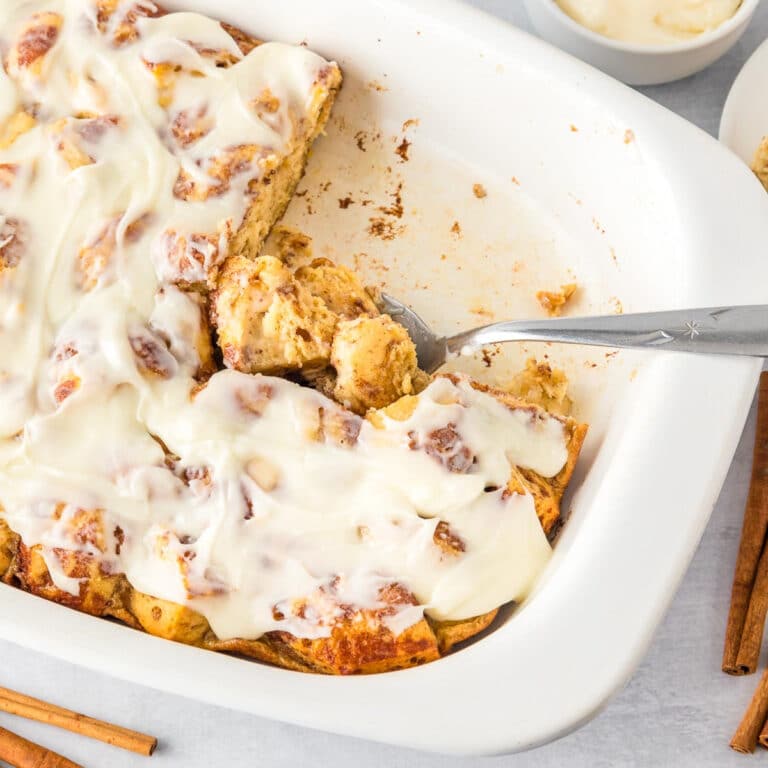 A white baking dish with frosted cinnamon roll casserole, partially served as a spoon scoops from the pan.