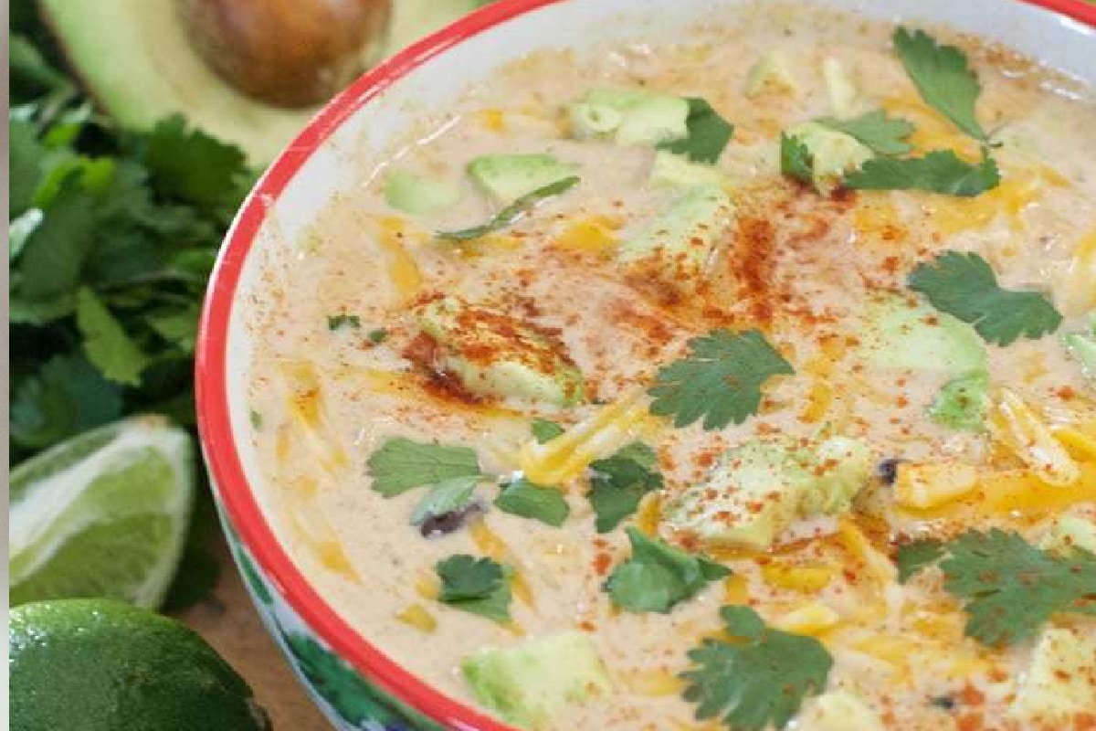 A bowl of creamy taco soup topped with diced avocado, shredded cheese, cilantro, and a sprinkle of paprika. Lime wedges and half an avocado are nearby.