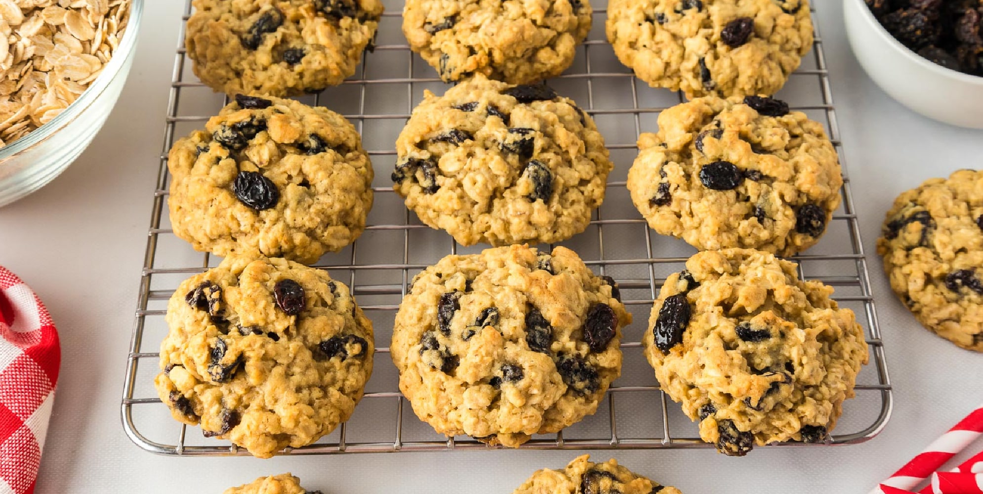 Wide view of oatmeal raisin cookies cooling on a wire rack with oats and raisins in bowls nearby.