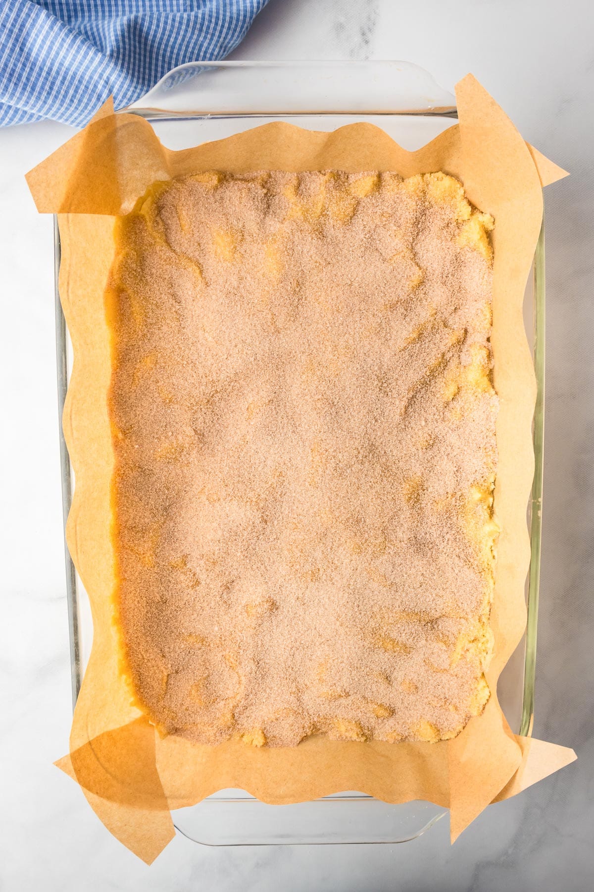 A glass baking dish lined with parchment paper full of snickerdoodle cookie bar dough topped with cinnamon and sugar before baking.