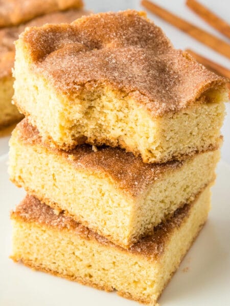 Three squares of snickerdoodle cookie bars stacked high on a plate with the top cookie bar missing a bite, with more cookie bars in the background.