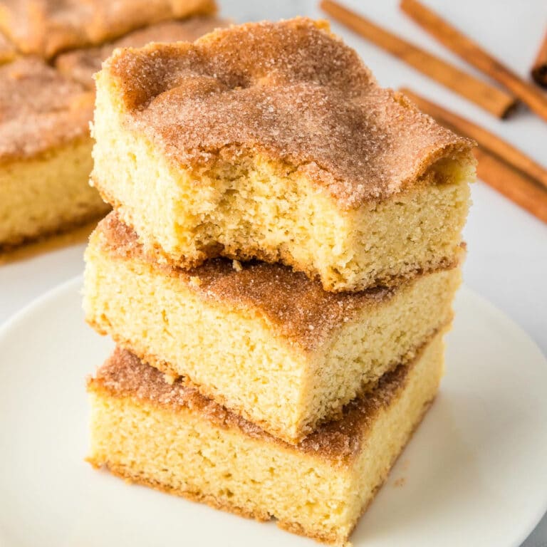 Three squares of snickerdoodle cookie bars stacked high on a plate with the top cookie bar missing a bite, with more cookie bars in the background.