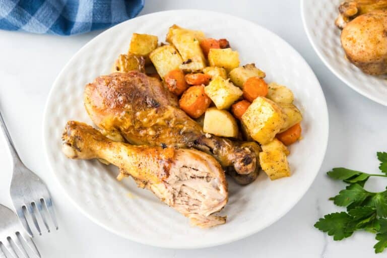 Wide view of a plate with roasted chicken drumsticks and seasoned potatoes and carrots with the chicken missing a bite.