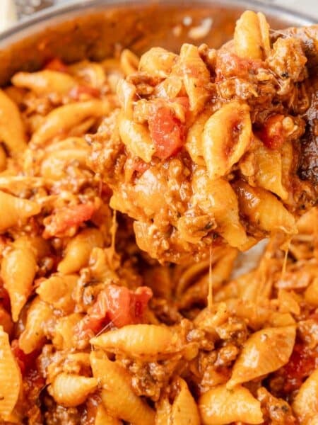 A wooden spoon scoops pasta shells mixed with ground meat, tomatoes, and melted cheese from a pan.
