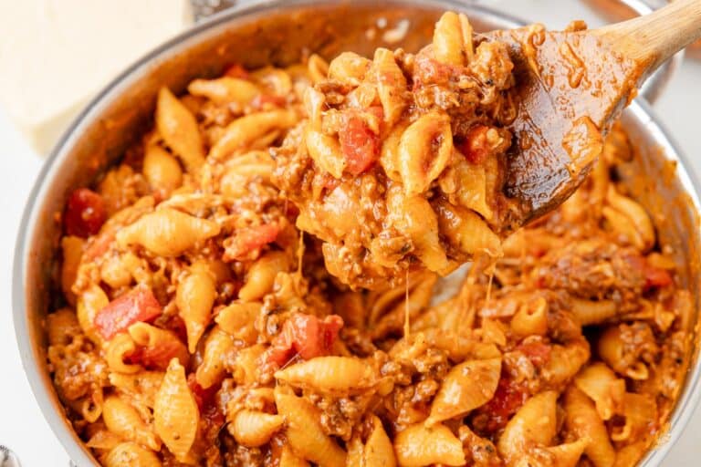 A wooden spoon scoops pasta shells mixed with ground meat, tomatoes, and melted cheese from a pan.