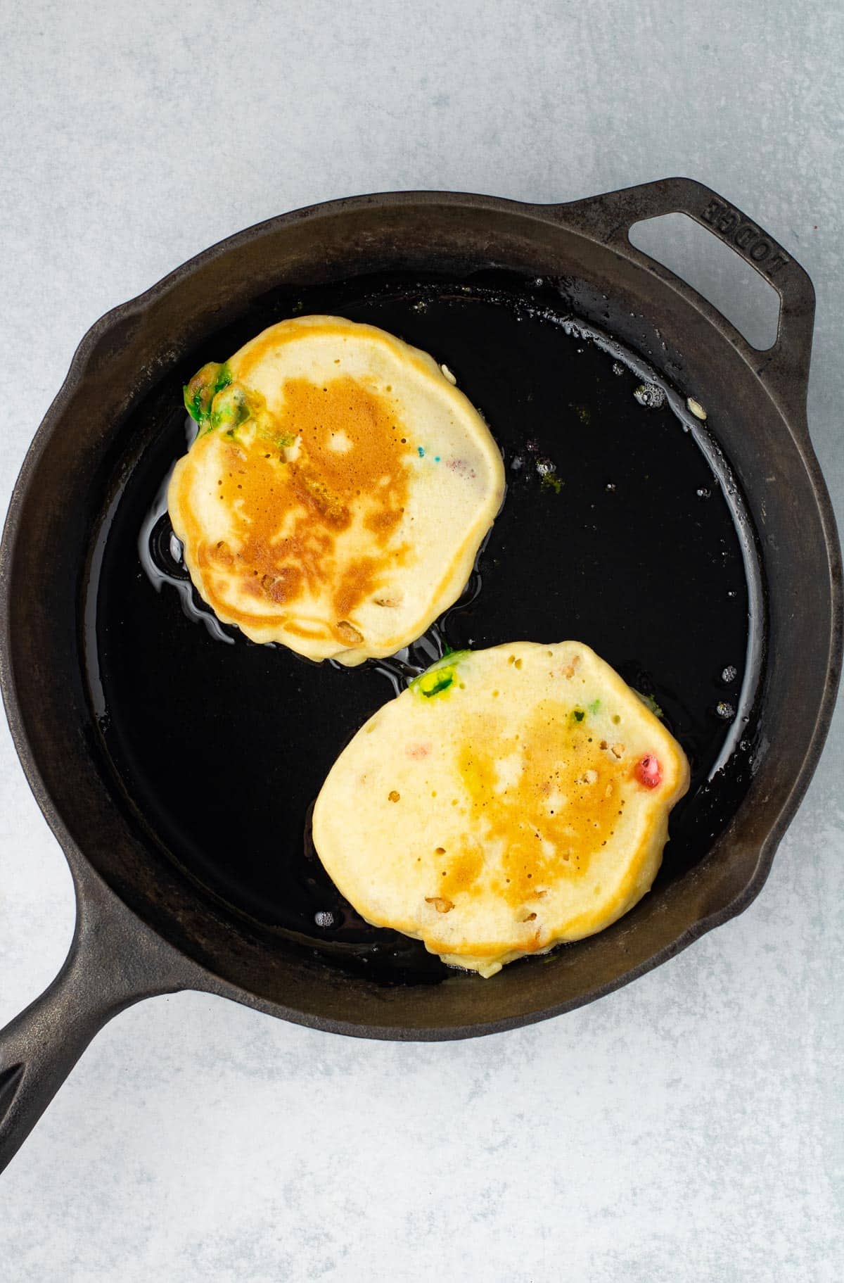 Two golden brown Lucky Charm pancakes cooking in a skillet.