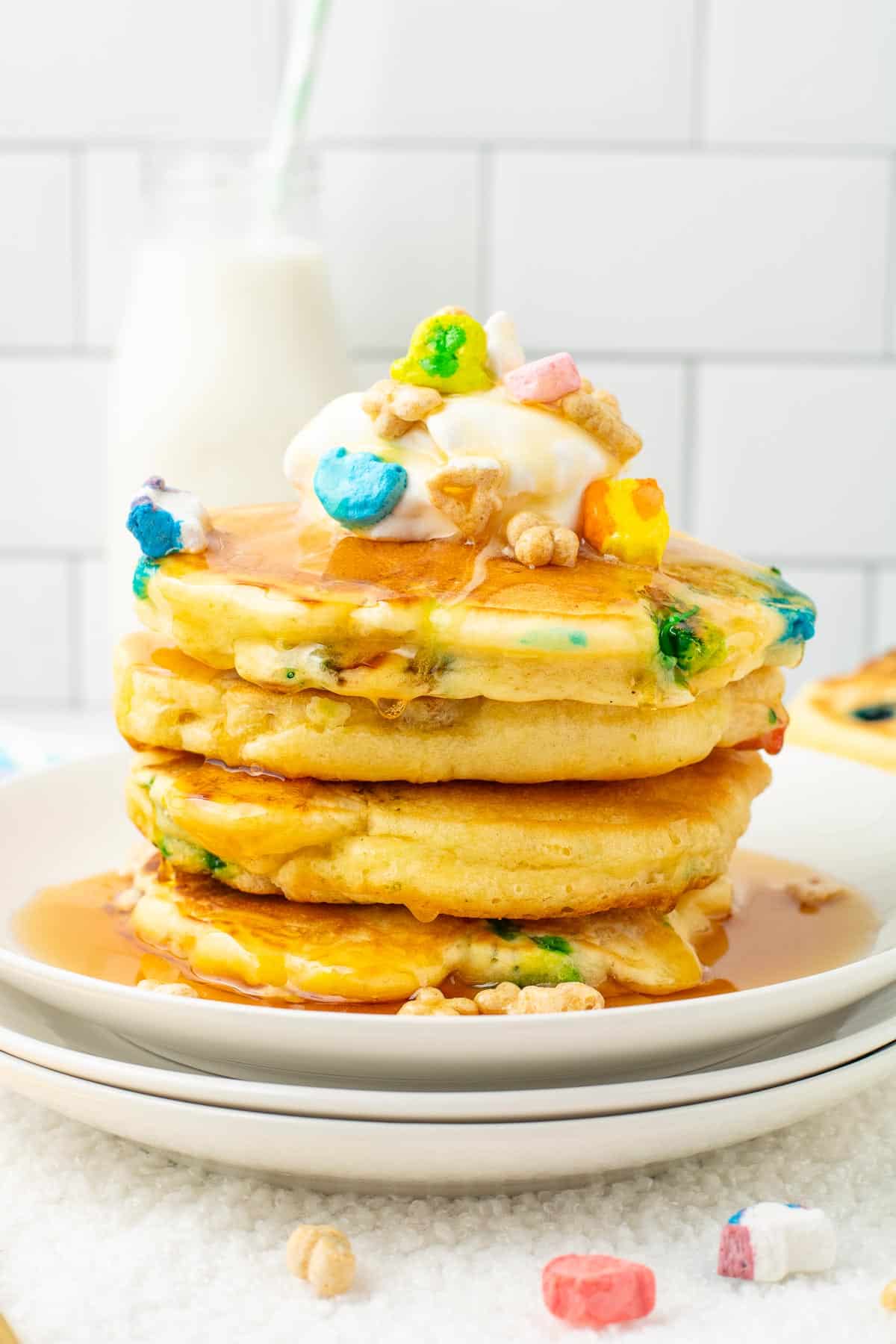 Stack of Lucky Charm pancakes topped with whipped cream, colorful cereal pieces, and syrup on a plate.