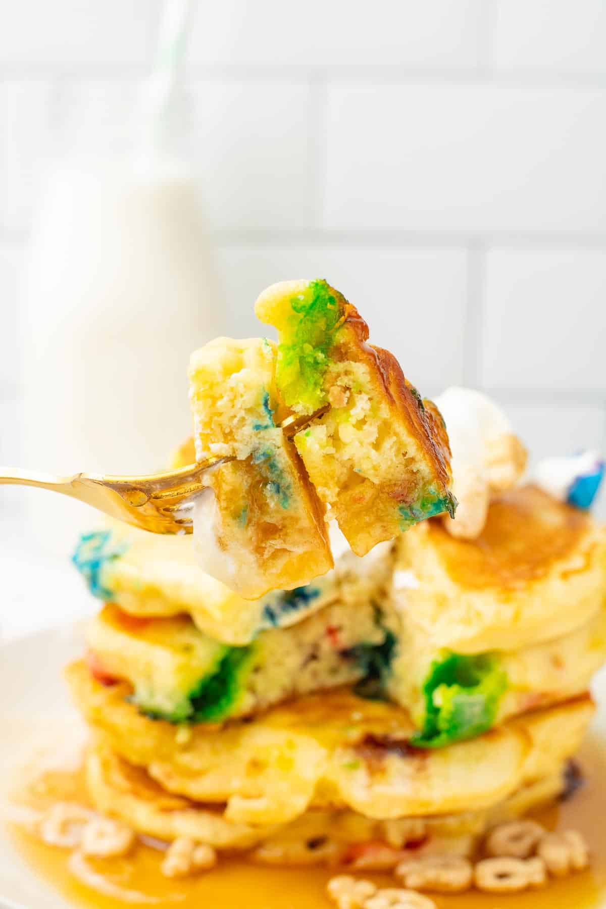 A fork holds a bite of colorful, Lucky Charm Pancakes in front of a stack.