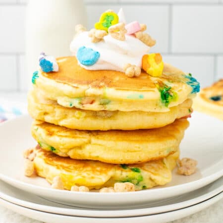 A square view of a stack of Lucky Charm pancakes topped with whipped cream on a plate.