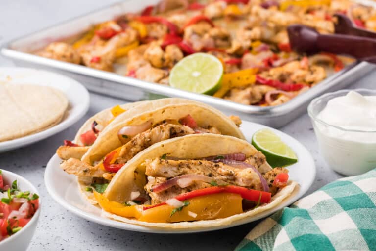 A plate of chicken fajitas with bell peppers and onions, accompanied by lime slices and sour cream. More fajitas are visible in a baking tray in the background.