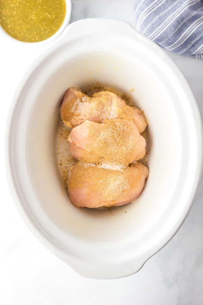 Raw chicken breasts seasoned with spices in a slow cooker base for salsa verde chicken.