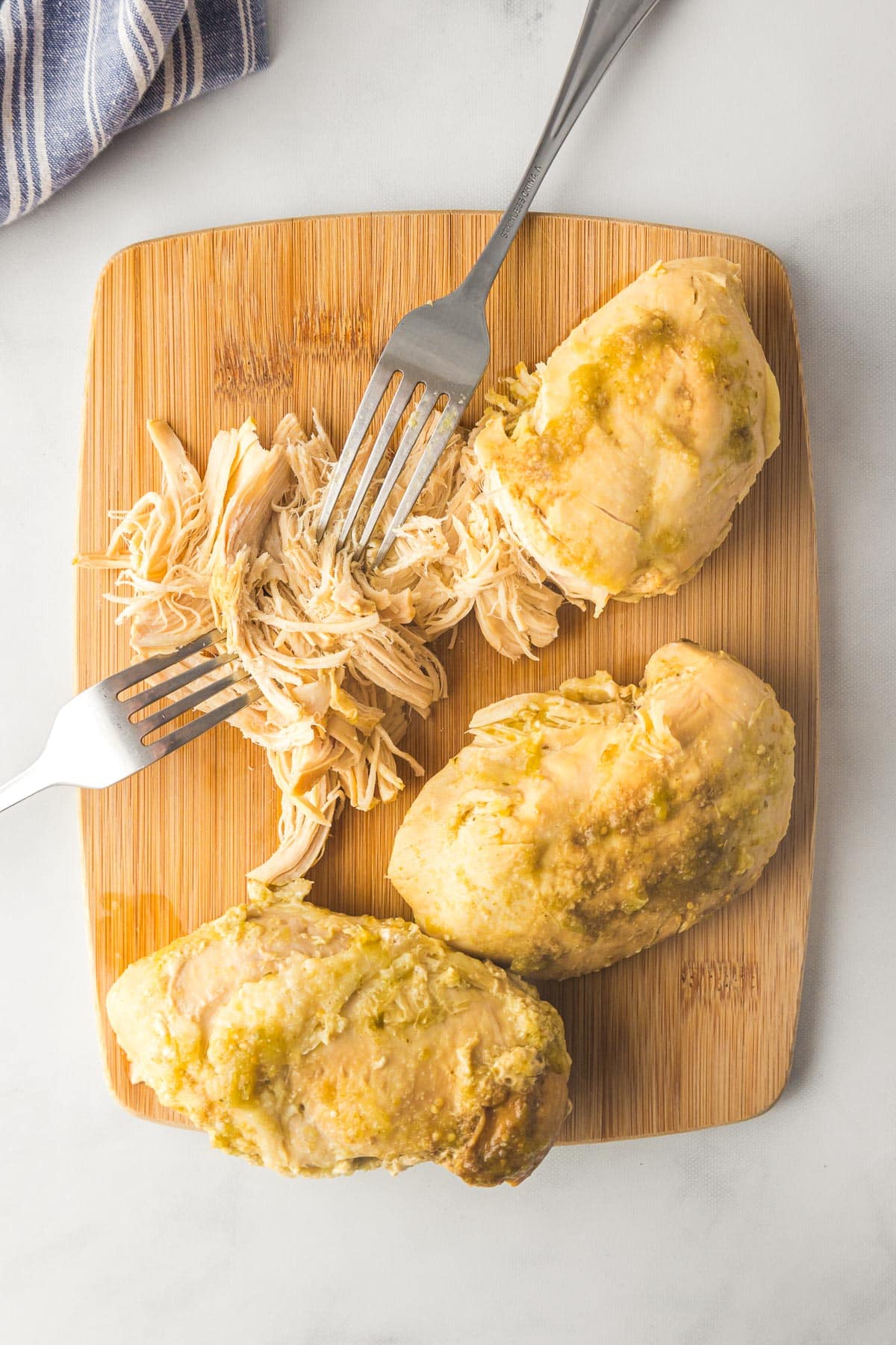 Three pieces of cooked chicken breast being shredded with two forks on a wooden cutting board for salsa verde chicken.