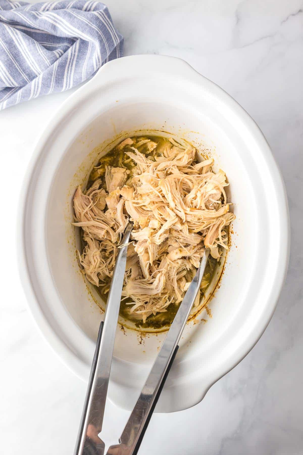 Shredded chicken being tossed in salsa verde in the base of a crockpot with metal tongs on a kitchen counter.