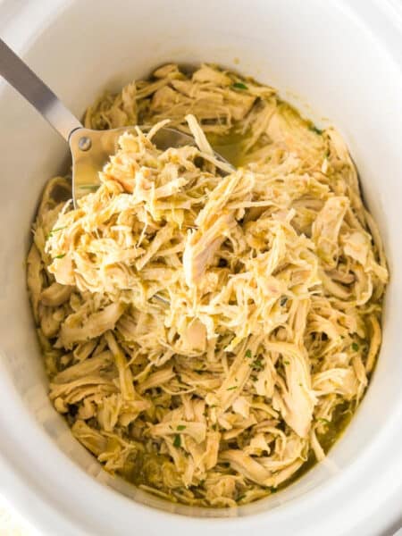 Square view of shredded salsa verde chicken in a slow cooker with a metal serving spoon scooping a spoonful.