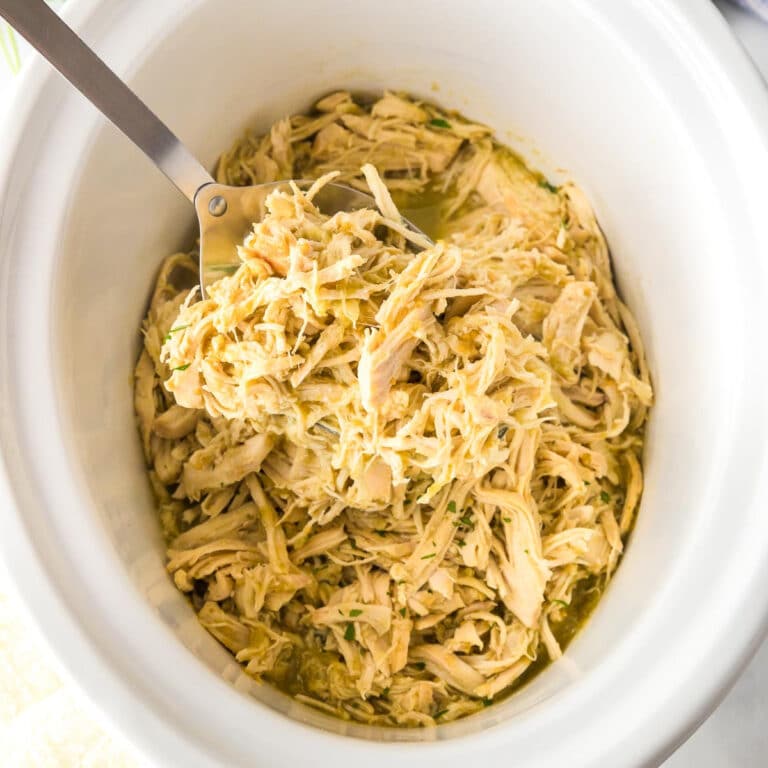 Square view of shredded salsa verde chicken in a slow cooker with a metal serving spoon scooping a spoonful.